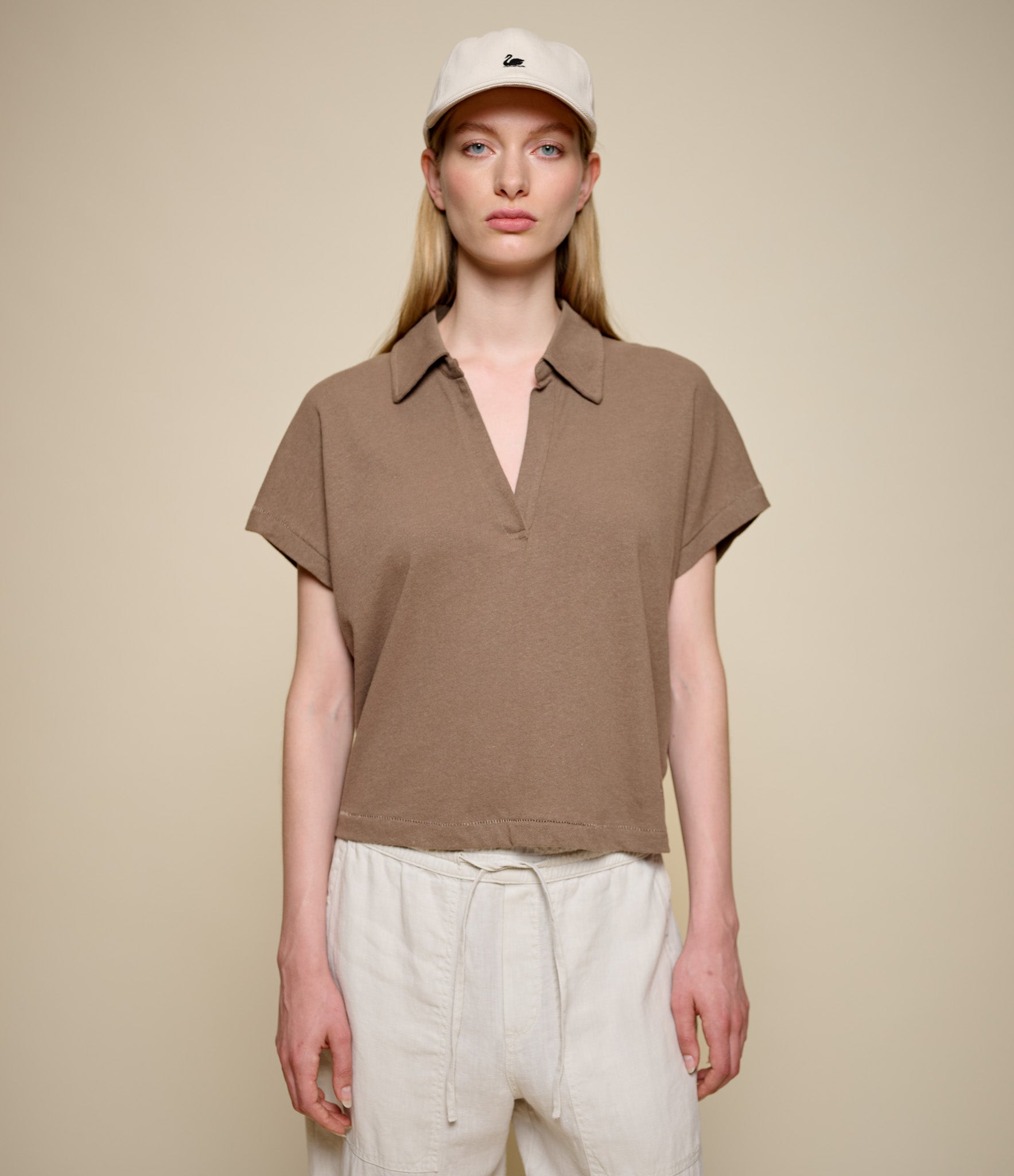 Woman wearing a brown linen polo shirt and beige cap against a beige background