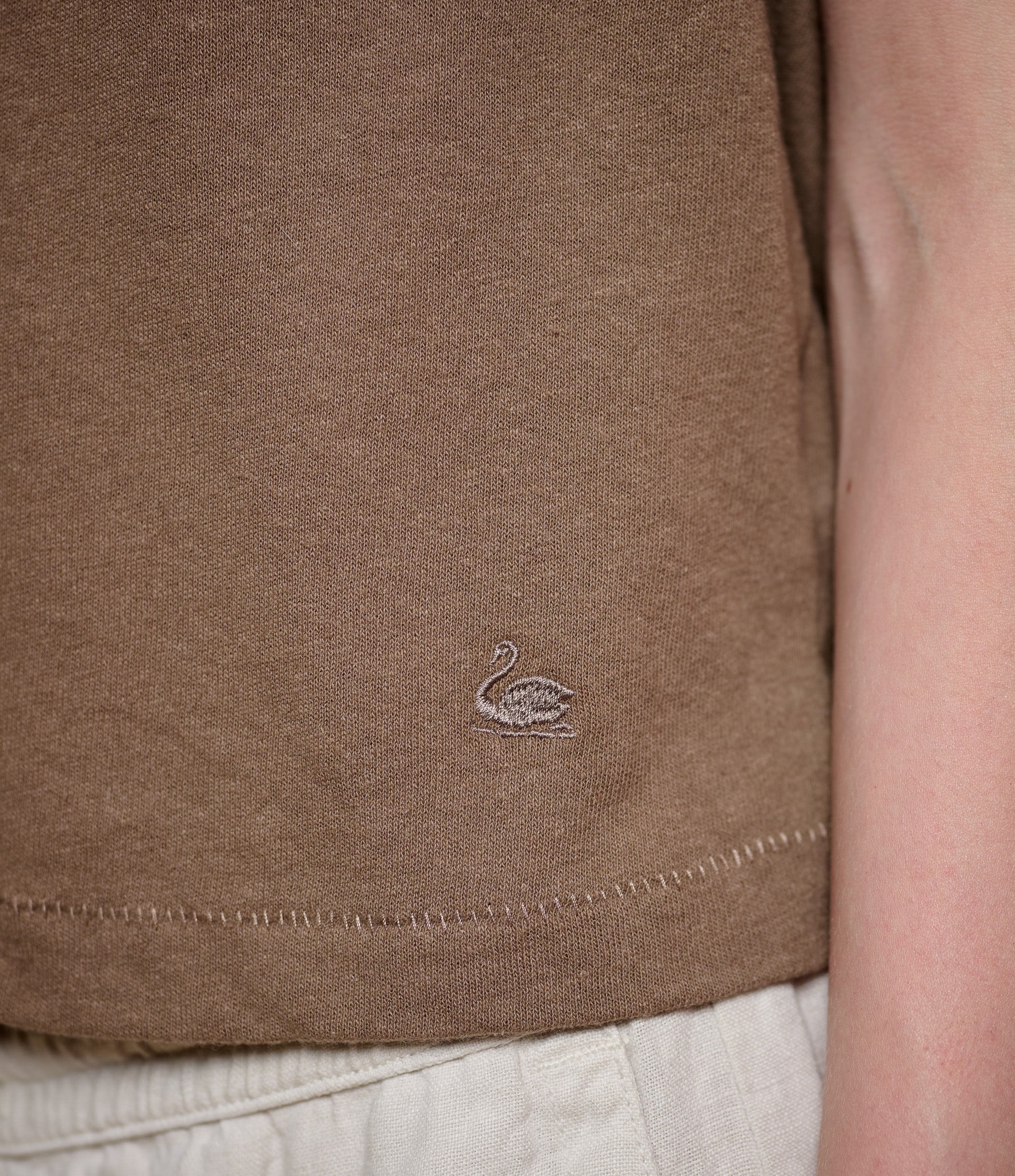 Close-up of brown polo shirt with a swan logo on a person wearing beige pants.