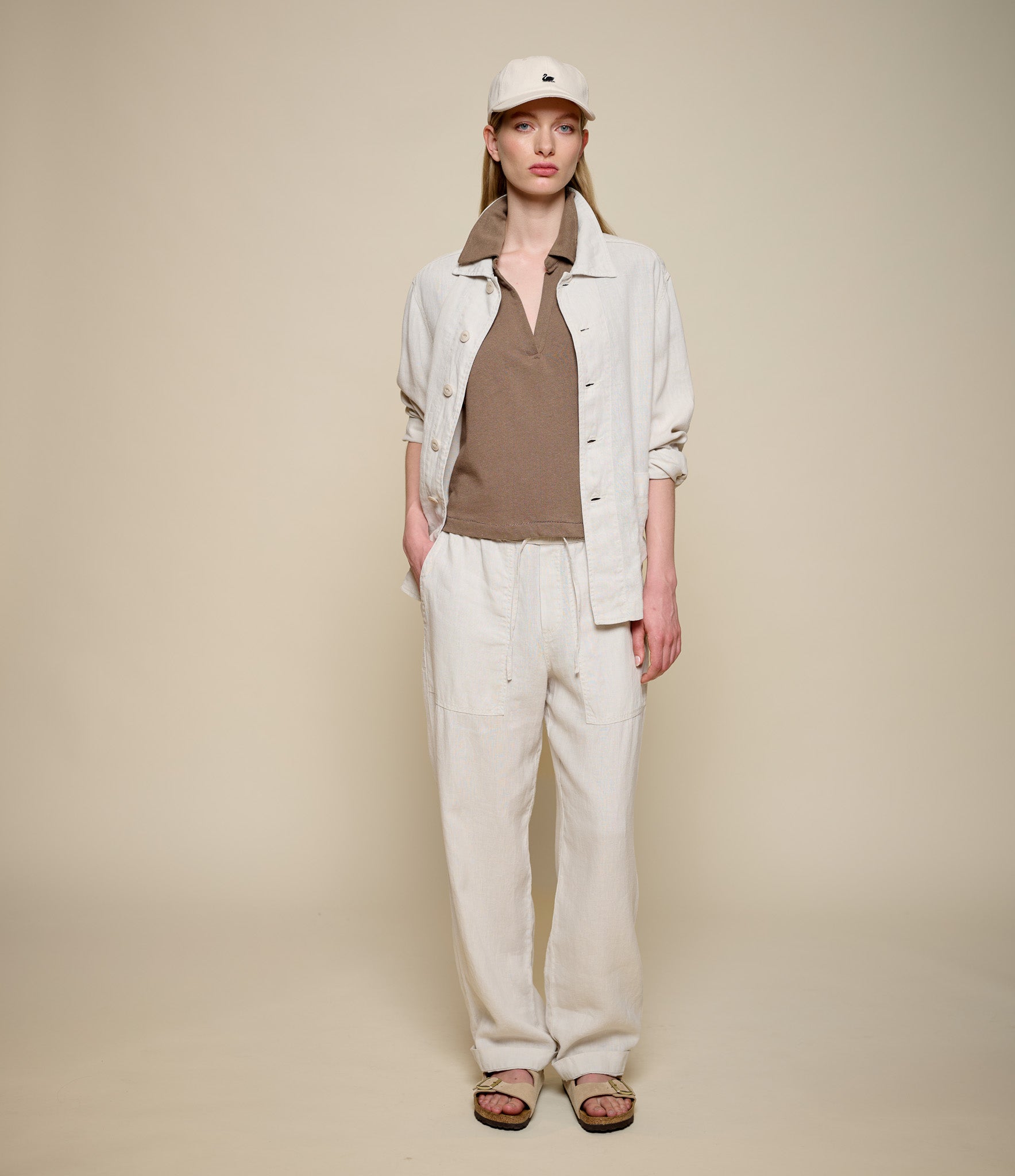 Woman wearing a beige linen jacket, brown linen polo shirt, and white linen pants against a beige background