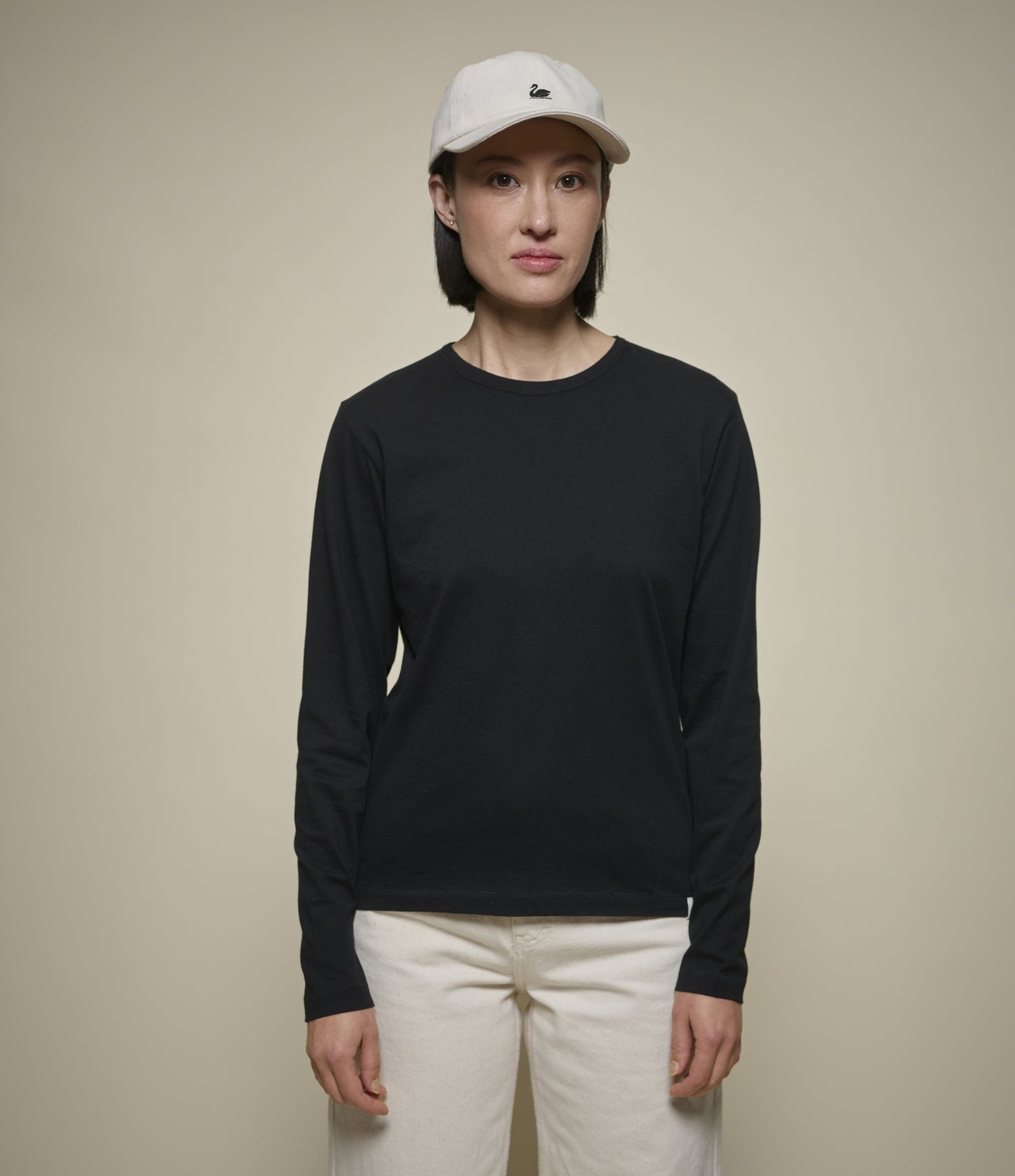 A woman modeling a black long-sleeve crewneck shirt, paired with off-white pants and a light beige baseball cap, against a solid beige background.