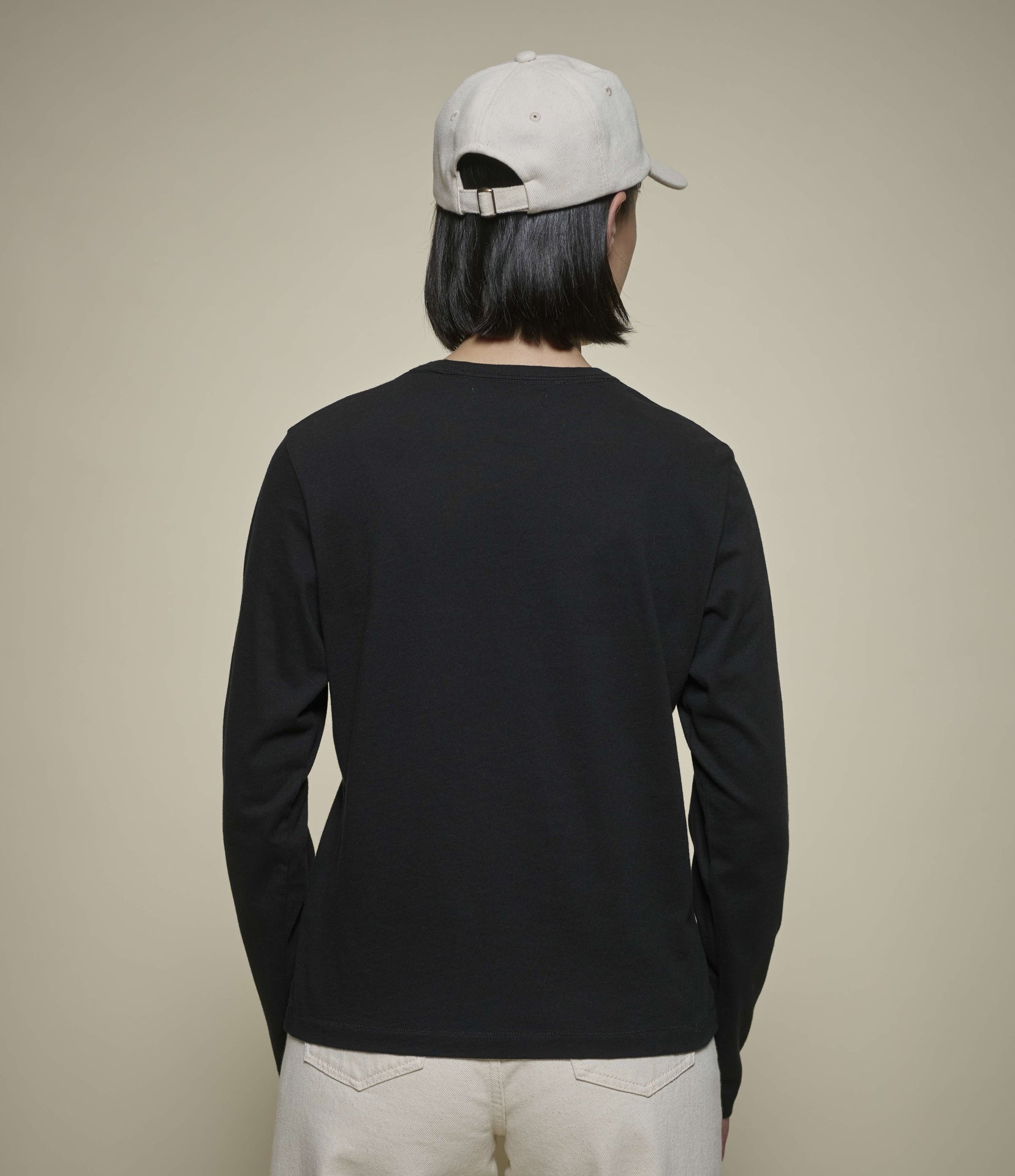 A person shown from the back wearing a black long-sleeve crewneck shirt, off-white pants, and a light gray baseball cap against a plain beige background.