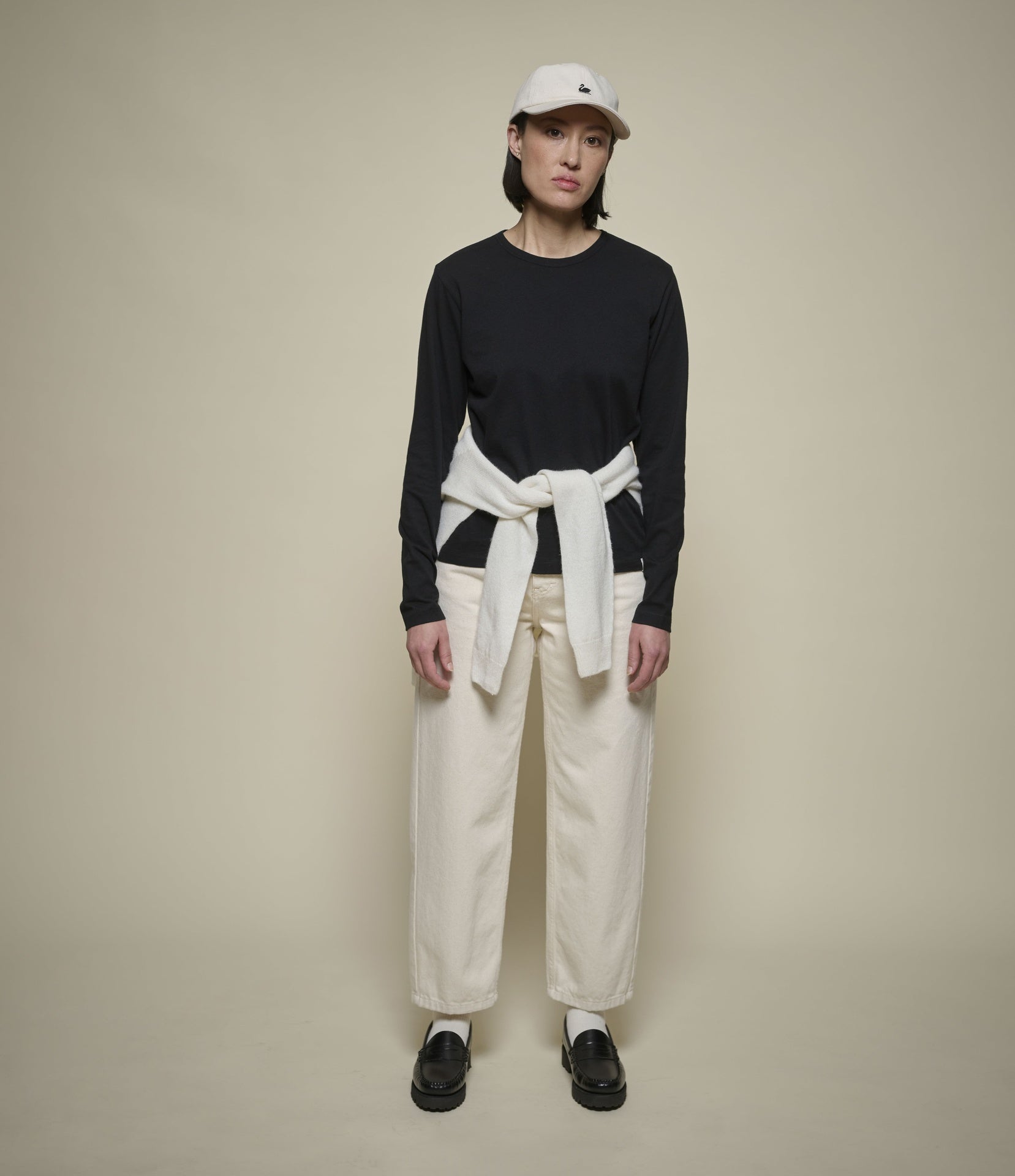 A model stands against a plain beige studio background wearing a black long-sleeve top, cream wide-leg pants, a cream shirt tied at the waist, black loafers, white socks, and a white cap.