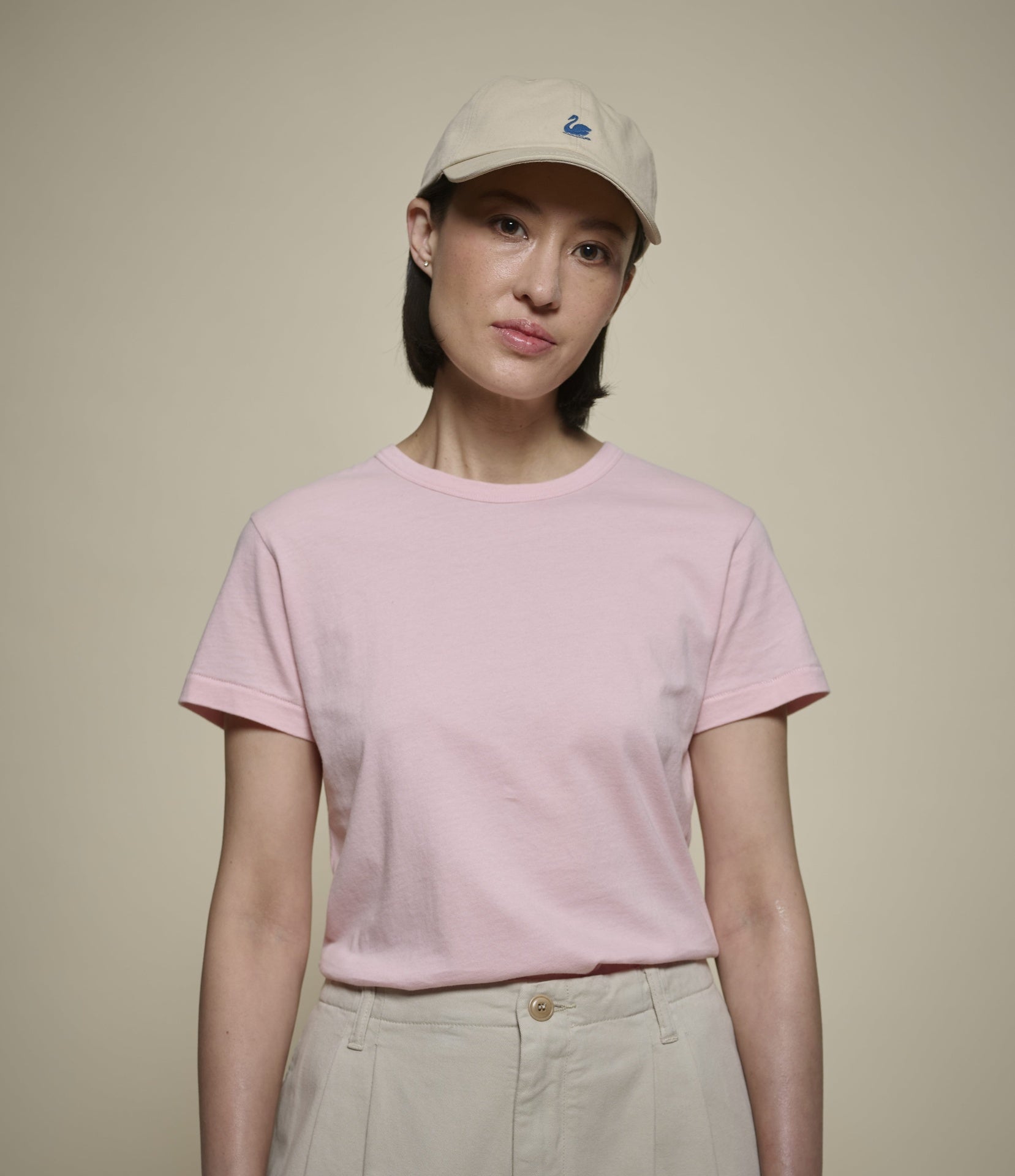 A woman wearing a light pink short-sleeve crewneck t-shirt, beige pleated trousers, and a beige baseball cap with a small blue logo, standing against a solid beige background.