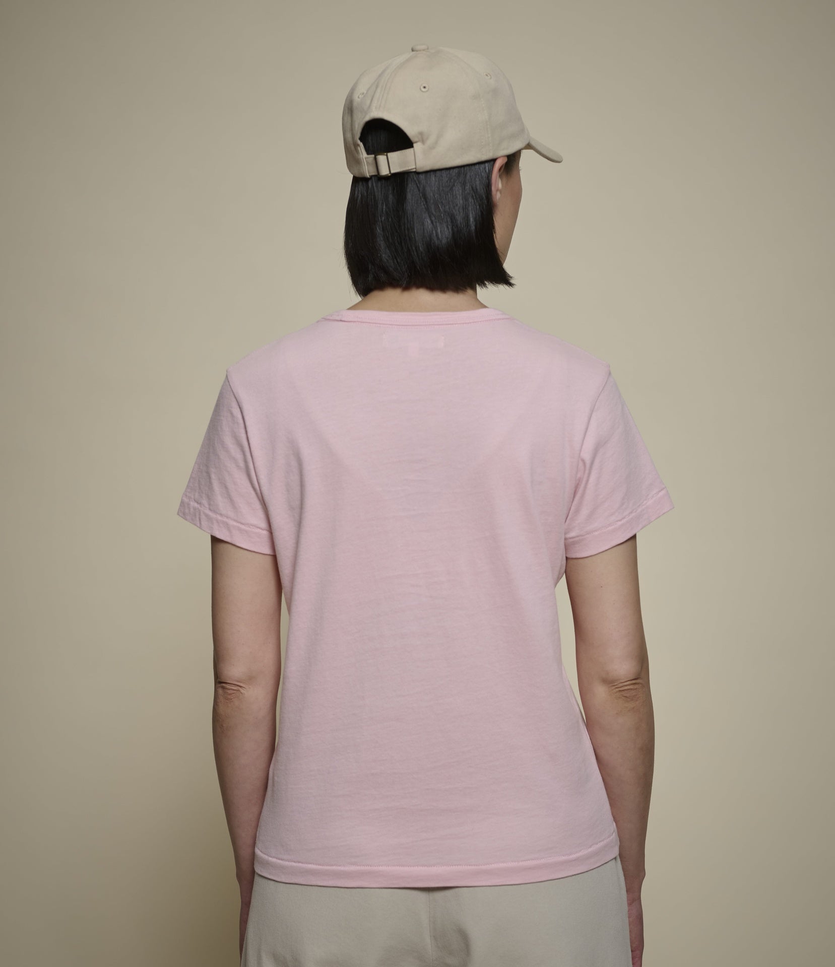 A woman shown from the back wearing a light pink short-sleeve crewneck t-shirt, a beige baseball cap, and beige pants, set against a solid beige background.