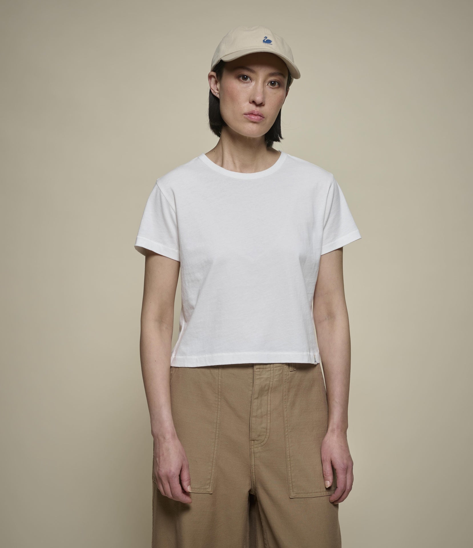 A model wearing a white short-sleeve cropped t-shirt, beige cargo pants, and a beige baseball cap, standing against a solid beige background.