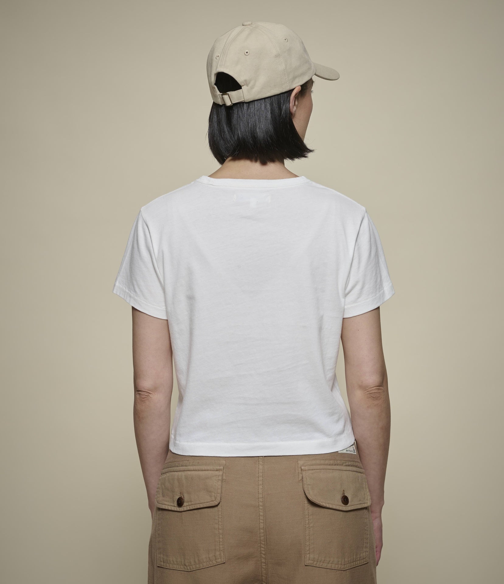 A person shown from the back wearing a plain white short-sleeve cropped T-shirt, tan pants, and a beige baseball cap against a plain beige studio background.