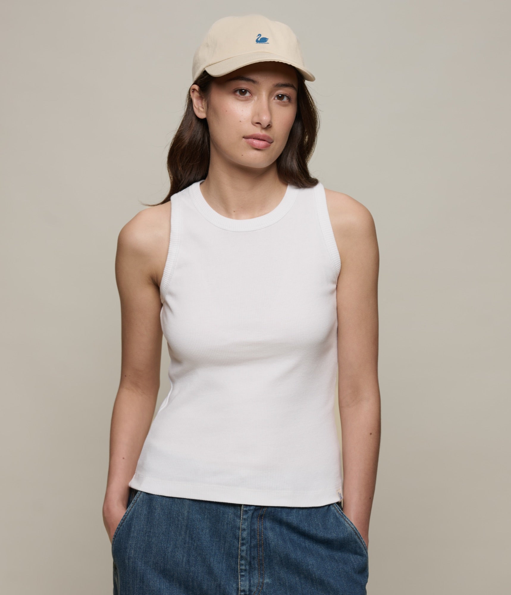 Person wearing a white ribbed tank top and blue jeans with a beige cap on a plain background