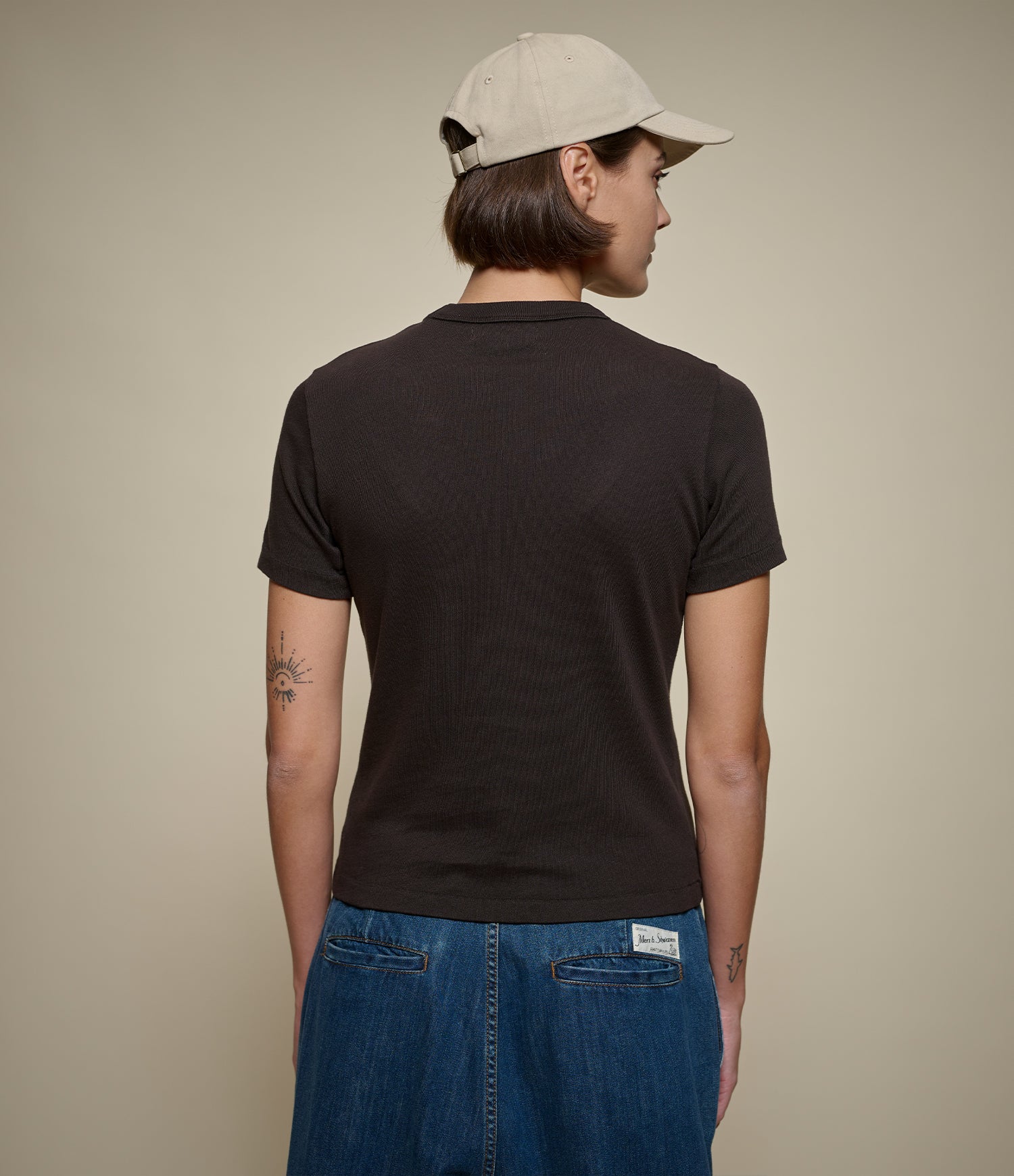 Woman shown from the back wearing a dark brown short-sleeve t-shirt, blue jeans, and a light beige baseball cap against a plain beige studio background.