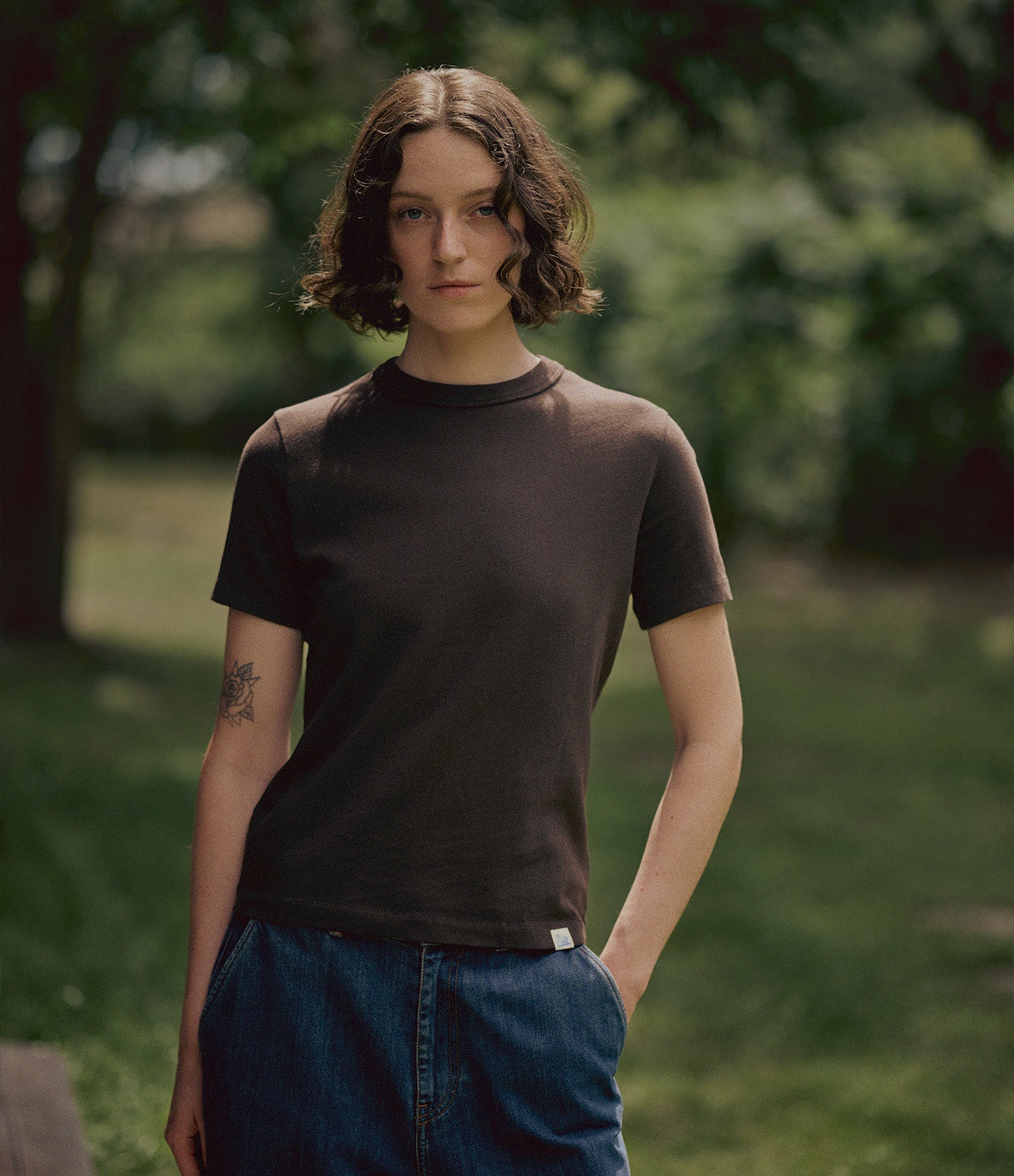 A woman wearing a dark brown short-sleeve crewneck t-shirt and blue jeans, standing outdoors in a grassy park area with trees in the background.