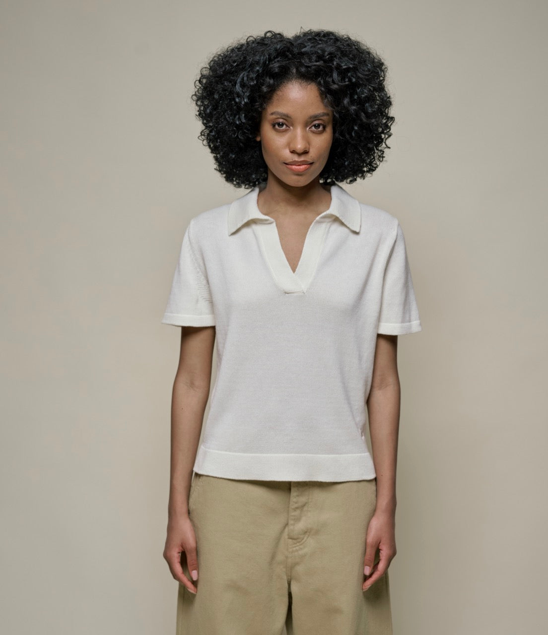 A person standing against a plain background, wearing a white knitted V-neck sweater with short sleeves and beige pants.