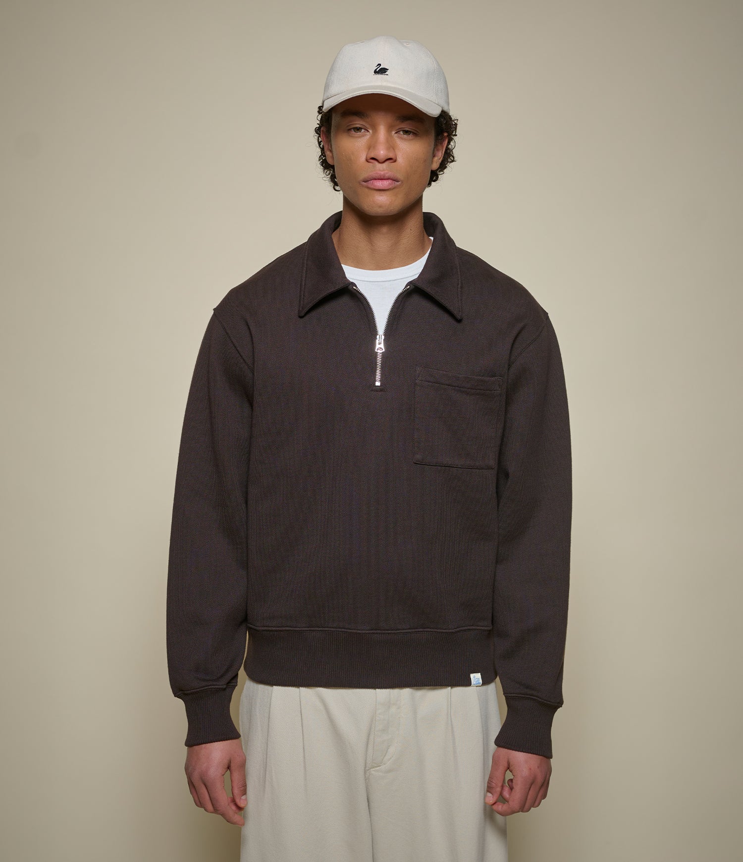 Man wearing a brown half-zip sweatshirt and beige cap against a beige background