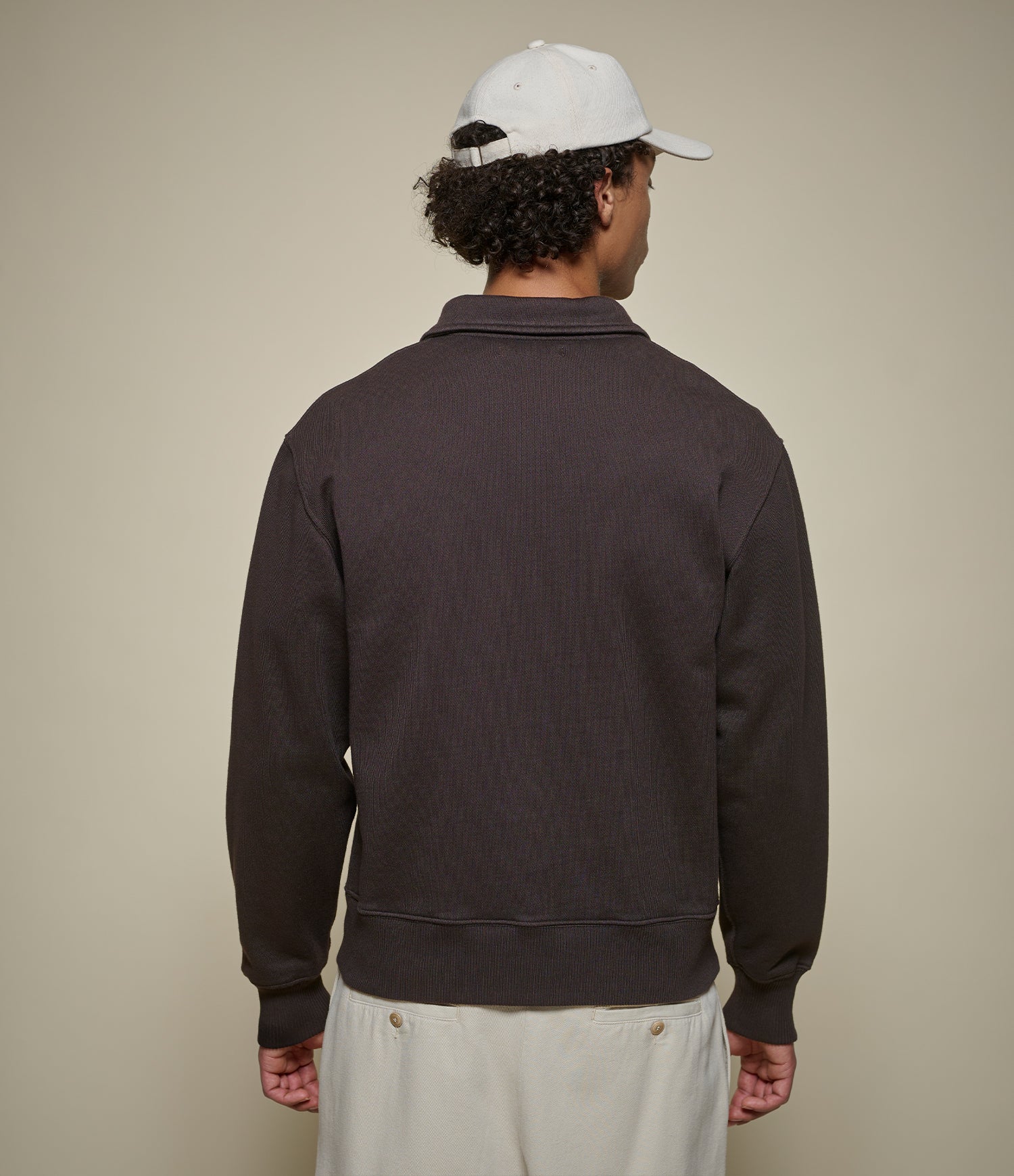Person wearing a dark brown sweatshirt and white cap from behind on a beige background