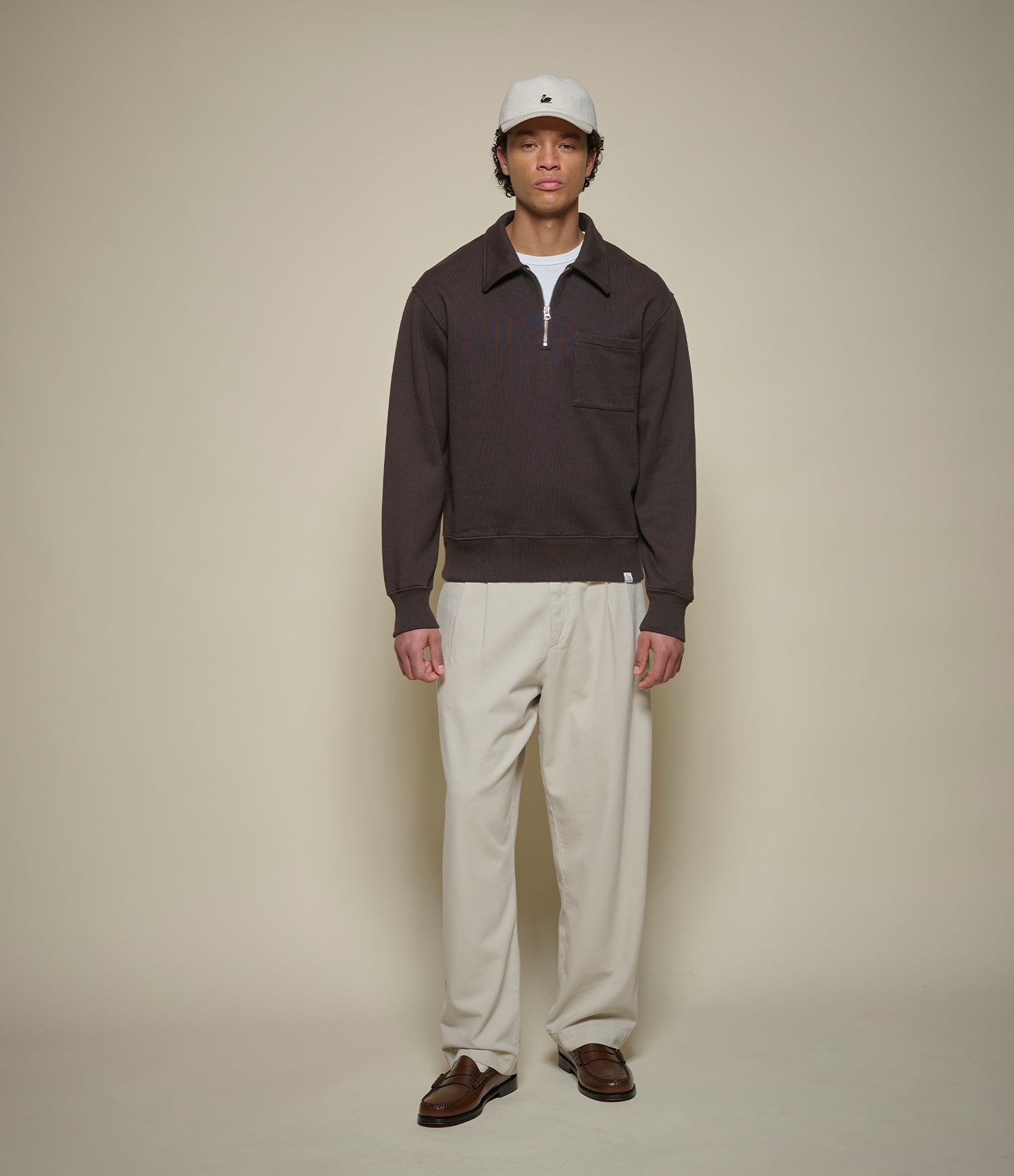 Person wearing a dark brown sweatshirt, white pants, and a white cap against a beige background