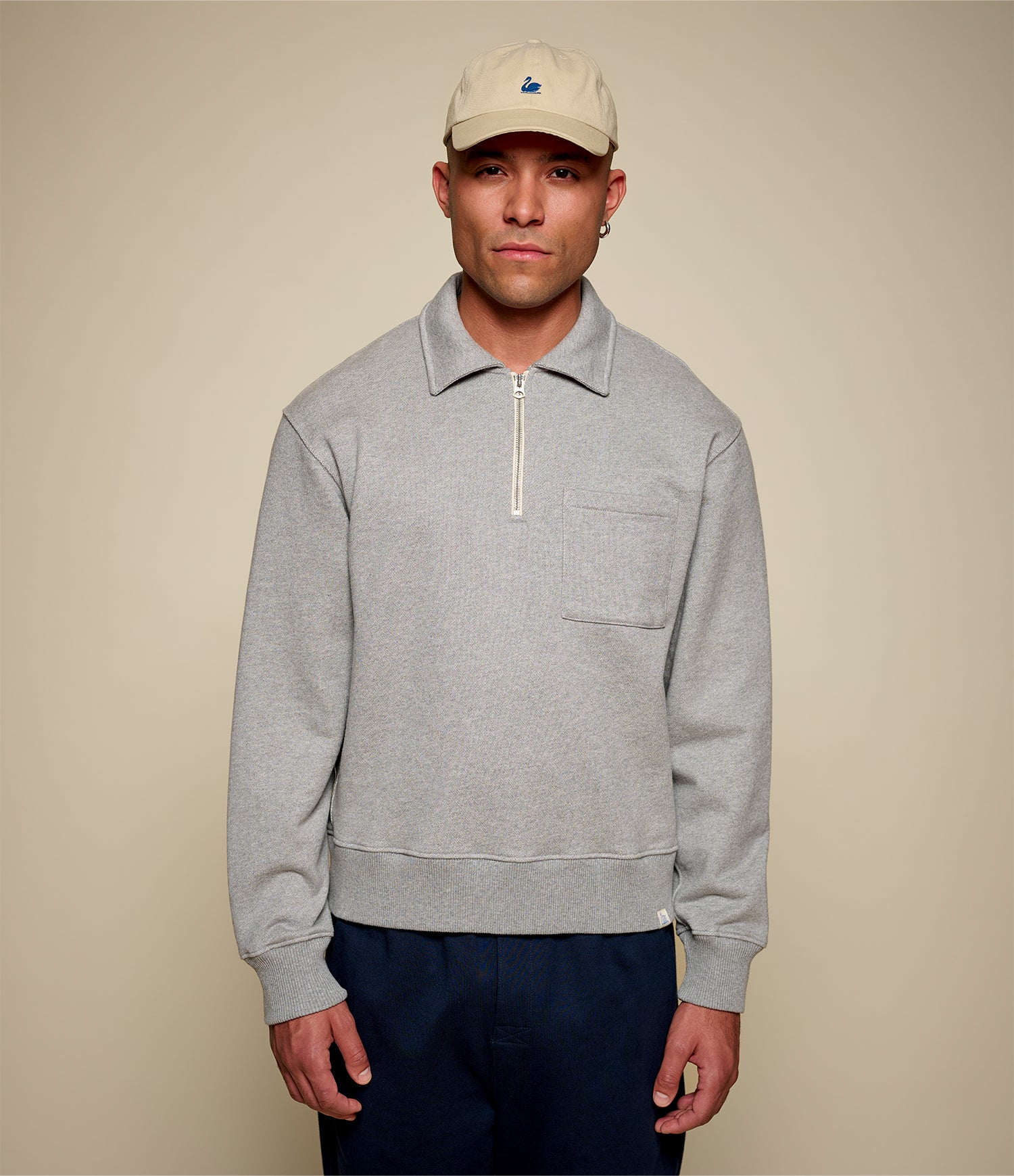 Man wearing a gray sweater with a zipper and a beige cap on a beige background
